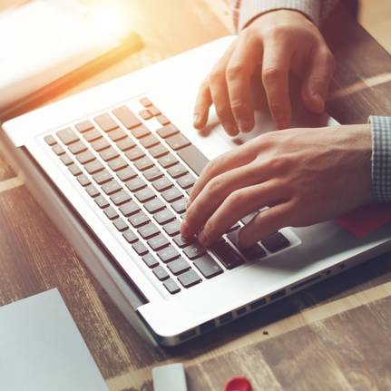 Das Bild zeigt eine Person, die an einem Laptop arbeitet. Auf dem Holztisch liegen Stifte, Papier und weitere Arbeitsutensilien. Sonnenlicht f&auml;llt sanft auf die Szene.