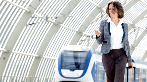 Frau läuft an Gleisen am Bahnhof entlang und schiebt ein Koffer währendd im Hintergrund ein Zug einfährt